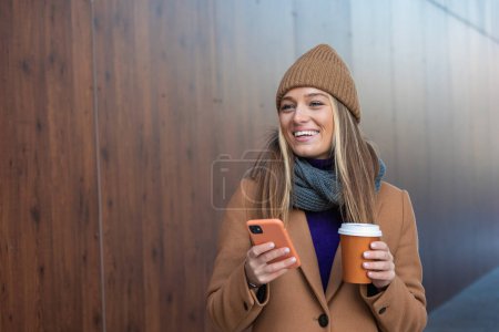 Foto de Hermosa mujer va a trabajar con café caminando cerca del edificio de oficinas. Retrato de una exitosa mujer de negocios sosteniendo una taza de bebida caliente. - Imagen libre de derechos