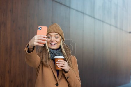 Téléchargez les photos : Jeune femme avec smartphone debout sur fond de bâtiment flou rue. Photo de mode de belle fille avec téléphone et tasse de café - en image libre de droit