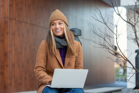 Téléchargez les photos : Belle jeune femme portant manteau d'automne en utilisant un ordinateur portable tout en étant assis sur un banc, boire tasse de café à emporter - en image libre de droit