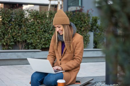 Photo for Attractive Caucasian girl working on a city street on a bench she holds a laptop in her hands, behind her a modern building, office worker, freelancer. - Royalty Free Image