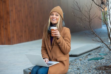Foto de Hermosa mujer joven bebiendo taza de café afuera en la cafetería de la calle trabajando en el ordenador portátil - Imagen libre de derechos