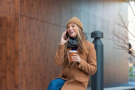 Foto de Blonde woman chatting, browsing internet online on smartphone during coffee break while relaxing on park bench. Beautiful cute young woman using mobile phone and holding cup of coffee. - Imagen libre de derechos