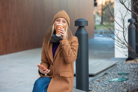 Foto de Blonde woman chatting, browsing internet online on smartphone during coffee break while relaxing on park bench. Beautiful cute young woman using mobile phone and holding cup of coffee. - Imagen libre de derechos
