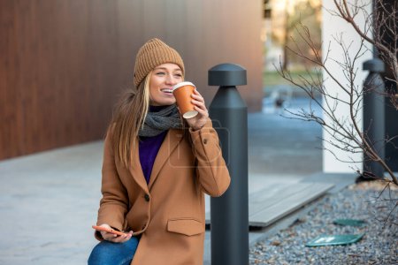 Foto de Retrato, Mujer joven atractiva y encantadora con ropa casual sentada en el banco, disfrutando de su café y usando su teléfono inteligente. - Imagen libre de derechos