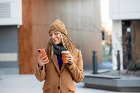 Téléchargez les photos : Jeune femme avec smartphone debout sur fond de bâtiment flou rue. Photo d'affaires de mode de belle fille avec téléphone et tasse de café - en image libre de droit