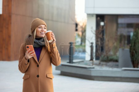 Téléchargez les photos : Jeune femme avec smartphone debout sur fond de bâtiment flou rue. Photo d'affaires de mode de belle fille avec téléphone et tasse de café - en image libre de droit