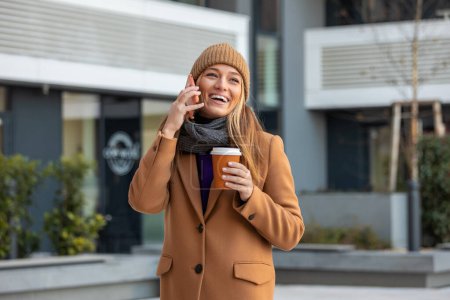 Téléchargez les photos : Young woman with smartphone standing against street blurred building background. Fashion business photo of beautiful girl with phone and cup of coffee - en image libre de droit