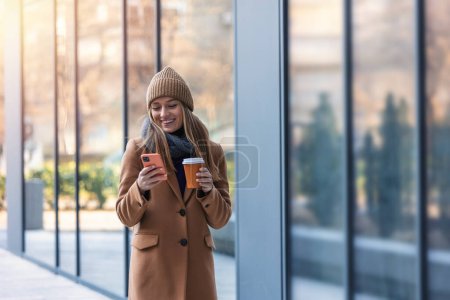 Photo for Cheerful young woman wearing coat walking outdoors, holding takeaway coffee cup, using mobile phone - Royalty Free Image