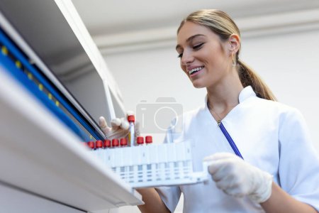 Foto de Retrato de una joven asistente de laboratorio haciendo análisis con tubos de ensayo y máquinas analizadoras sentadas en el moderno laboratorio - Imagen libre de derechos