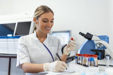 Photo for Closeup portrait of woman scientist working in lab. Female researcher sitting at desk looking through microscope at blood and chemical test sample and making notes in clipboard. Biochemistry work - Royalty Free Image