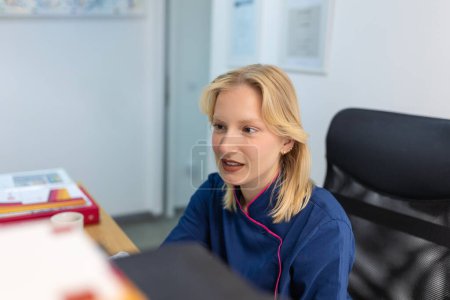 Téléchargez les photos : Une femme médecin en uniforme travaille avec un document dans une clinique privée. Infirmière assise derrière le bureau au bureau, vérifiant les antécédents médicaux ou l'anamnèse du client, lisant le plan de traitement du patient - en image libre de droit