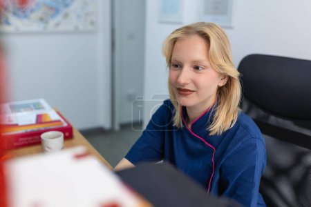 Téléchargez les photos : Une femme médecin en uniforme travaille avec un document dans une clinique privée. Infirmière assise derrière le bureau au bureau, vérifiant les antécédents médicaux ou l'anamnèse du client, lisant le plan de traitement du patient - en image libre de droit
