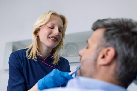 Téléchargez les photos : L'homme est venu voir le dentiste. Il est assis sur la chaise dentaire. Le dentiste s'est penché sur lui. Concept patient heureux et dentiste. - en image libre de droit