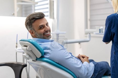 Téléchargez les photos : Image de l'homme satisfait assis dans une chaise dentaire au centre médical tandis que le médecin professionnel fixant ses dents - en image libre de droit