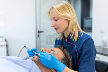 Foto de Dentista en uniforme azul haciendo procedimiento de dientes al paciente, paciente masculino durante el procedimiento de tratamiento en la clínica dental contemporánea - Imagen libre de derechos