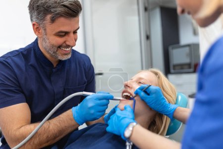 Photo for Dentist in blue uniform with assistant doing teeth procedure to patient female patient with turbine during treatment procedure in contemporary dental clinic - Royalty Free Image