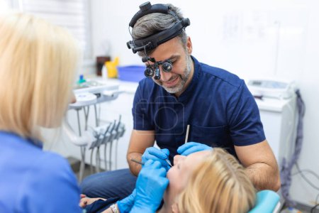 Photo for Man dentist and his assistants doing treatment for patient blonde lady, holding dental tools, wearing rubber gloves. Stomatology, dentistry, modern dental clinic concept - Royalty Free Image