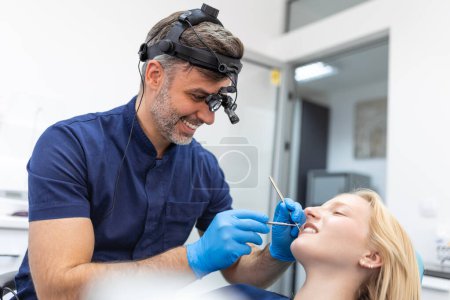Téléchargez les photos : Smiling brunette woman being examined by dentist at dental clinic. Hands of a doctor holding dental instruments near patient's mouth. Healthy teeth and medicine concept - en image libre de droit