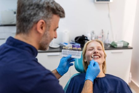 Photo for Image of satisfied young woman sitting in dental chair at medical center while professional doctor fixing her teeth, Female dentist choosing filling shade for smiling woman, using tooth scale sample - Royalty Free Image