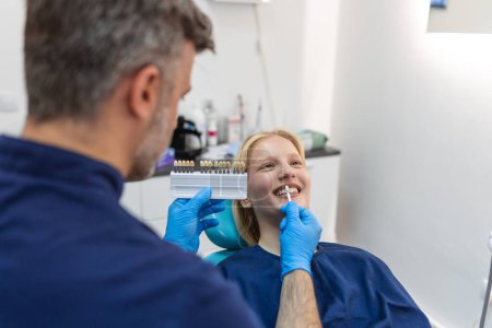 Téléchargez les photos : Image of satisfied young woman sitting in dental chair at medical center while professional doctor fixing her teeth, Female dentist choosing filling shade for smiling woman, using tooth scale sample - en image libre de droit