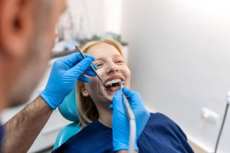 Téléchargez les photos : Dentiste souriant communiquant avec la jeune femme tout en vérifiant ses dents pendant la procédure dentaire au cabinet du dentiste. - en image libre de droit