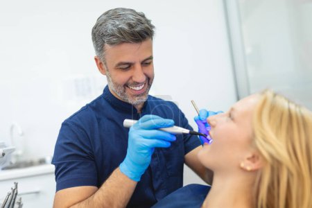 Téléchargez les photos : Jeune femme pendant la procédure de séchage de remplissage dentaire avec la lumière UV de durcissement à la clinique dentaire. - en image libre de droit