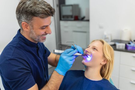 Téléchargez les photos : Jeune femme pendant la procédure de séchage de remplissage dentaire avec la lumière UV de durcissement à la clinique dentaire. - en image libre de droit