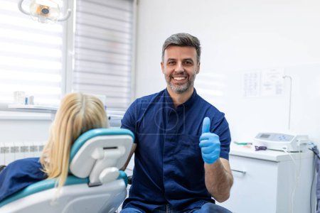 Foto de Hombre dentista posando en la clínica sobre gabinete moderno, espacio vacío. Retrato de un joven dentista sonriente sentado en su clínica mostrando signo OK - Imagen libre de derechos