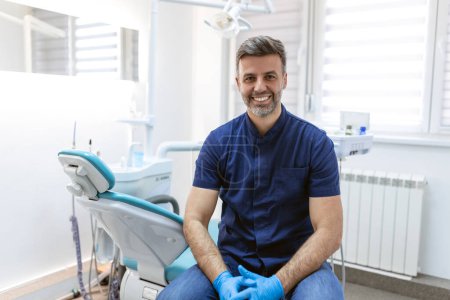Foto de Hombre dentista sonriendo mientras está parado en la clínica dental. Retrato de un joven dentista confiado que trabaja en su consultorio - Imagen libre de derechos