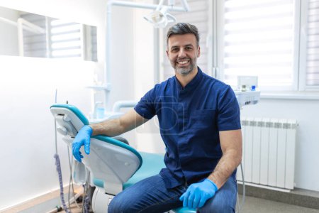 Photo for Male dentist posing at clinic over modern cabinet, empty space. Portrait shot of a young smiling dentist sitting in his clinic - Royalty Free Image