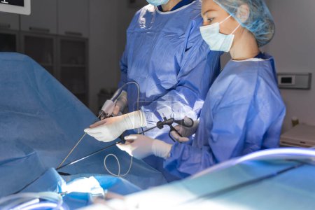 Téléchargez les photos : Vue arrière de l'équipe de chirurgiens examine les moniteurs tout en préformant l'opération dans le bloc opératoire de l'hôpital, chirurgien homme opérant patient travaillant avec des instruments de laparoscopie chirurgicale. - en image libre de droit