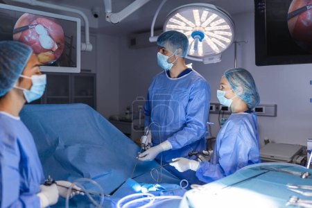 Foto de Vista posterior del equipo de cirujanos observa los monitores mientras se realiza la operación de preformación en el quirófano del hospital, paciente operado por cirujano masculino que trabaja con instrumentos de laparoscopia quirúrgica. - Imagen libre de derechos