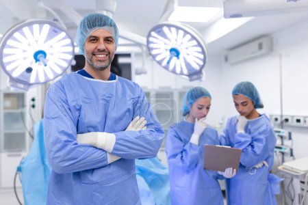 Téléchargez les photos : Portrait d'un chirurgien homme debout les bras croisés en salle d'opération à l'hôpital. Les chirurgiens d'équipe effectuent une opération, médecin d'âge moyen regarde la caméra, dans une salle d'opération moderne - en image libre de droit