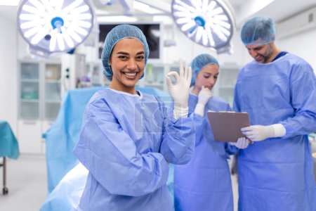 Foto de Close-up of a surgeon woman looking at camera with colleagues performing in background in operation room. The concept of medicine - Imagen libre de derechos