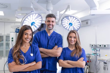 Téléchargez les photos : Groupe de trois médecins et infirmières debout dans un couloir d'hôpital, portant des blouses. L'équipe de travailleurs de la santé regarde la caméra et sourit - en image libre de droit