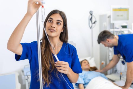 Foto de Mujer joven médico anestesiólogo vestido con bata azul, pone el gotero IV en la habitación del hospital - Imagen libre de derechos