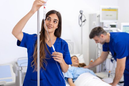 Foto de Mujer joven médico anestesiólogo vestido con bata azul, pone el gotero IV en la habitación del hospital - Imagen libre de derechos