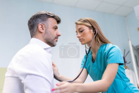 Téléchargez les photos : Le jeune médecin utilise un stéthoscope écouter le rythme cardiaque du patient. Prise de vue d'une femme médecin donnant un examen médical à un patient masculin - en image libre de droit