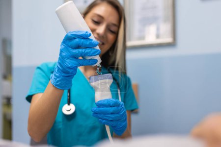 Foto de Médico sonriente que aplica gel en el dispositivo de ultrasonido antes del tratamiento. Las manos en los guantes aplican una tira de gel transparente al sensor ultrasónico del escáner. - Imagen libre de derechos