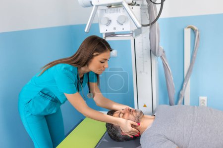 Foto de Paciente masculino acostado en la cama mientras enfermera ajustando la moderna máquina de rayos X para escanear su pecho en busca de lesiones y fracturas - Imagen libre de derechos