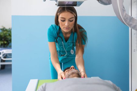 Téléchargez les photos : Patient homme couché sur le lit tandis que infirmière ajustant moderne appareil à rayons X pour scanner sa poitrine pour les blessures et les fractures - en image libre de droit