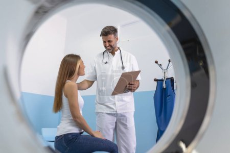 Foto de Técnico médico de pie y hablando con su paciente sentado en la cama del escáner CT. - Imagen libre de derechos