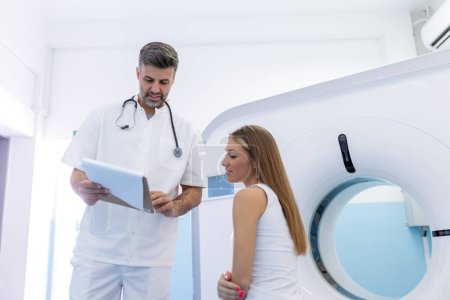 Téléchargez les photos : Jeune technicien médical parlant à son patient assis sur le lit du scanner CT
. - en image libre de droit