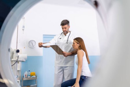 Téléchargez les photos : Jeune technicien médical parlant à son patient assis sur le lit du scanner CT
. - en image libre de droit