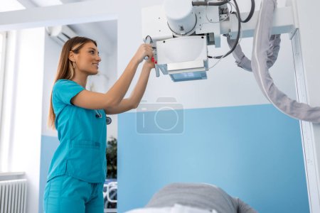 Foto de En el hospital, bajo ángulo de tiro del hombre acostado en una cama, Técnico femenino ajusta la máquina de rayos X. Escaneo de fracturas, extremidades rotas, lesiones, cáncer o tumor. Hospital moderno - Imagen libre de derechos
