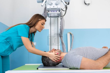 Foto de Una joven radióloga doctora confiada, tomando rayos X de un paciente varón acostado en la mesa de la máquina. Médico de pie cerca del paciente durante el procedimiento de rayos X de cabeza - Imagen libre de derechos