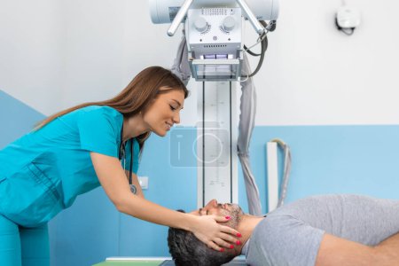 Foto de Una joven radióloga doctora confiada, tomando rayos X de un paciente varón acostado en la mesa de la máquina. Médico de pie cerca del paciente durante el procedimiento de rayos X de cabeza - Imagen libre de derechos