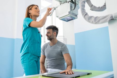 Téléchargez les photos : Patient masculin couché sur le lit pendant que l'infirmière ajustant la machine à rayons X moderne pour scanner son bras ou sa main pour les blessures et les fractures - en image libre de droit