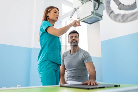 Téléchargez les photos : Patient masculin assis pendant que l'infirmière ajustant la machine à rayons X moderne pour scanner son bras ou sa main pour les blessures et les fractures - en image libre de droit