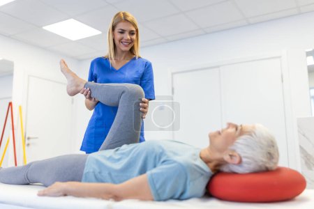 Téléchargez les photos : Gros plan de physiothérapeute féminine aide la femme aînée aînée étirant son string et faisant de la rééducation des cuisses ou des jambes dans la salle d'exercice - elle est allongée sur un lit de massage - en image libre de droit
